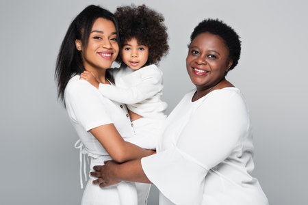 pleased african american women in white clothes looking at camera while holding child isolated on greyの写真素材