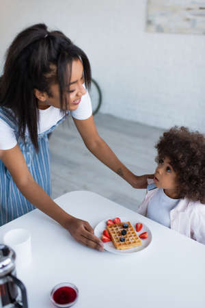 african american girl looking at smiling mother serving delicious waffle with fresh berries for breakfastの写真素材