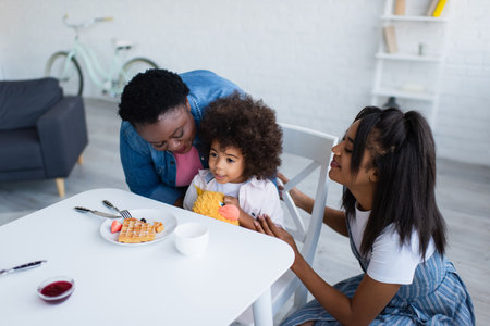 happy african american women hugging child sitting near waffle with berries and jam on tableの写真素材