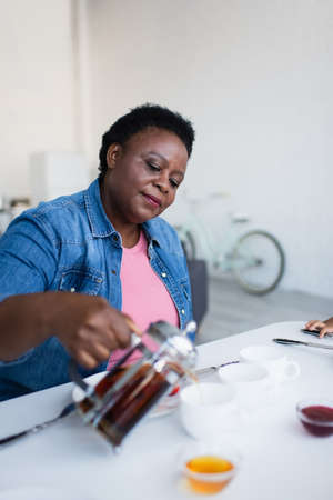 mature african american woman pouring tea near blurred bowls with jam and honey on tableの写真素材