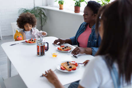 african american girl having breakfast with granny and blurred mom while sitting at table with soft toyの写真素材