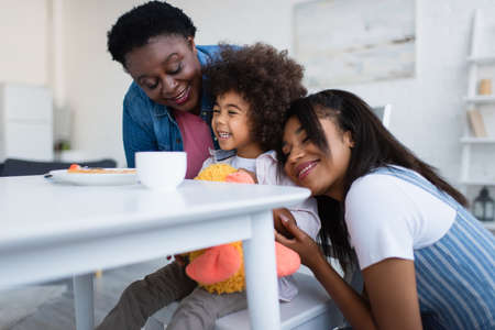 happy african american women hugging cheerful girl during breakfastの写真素材