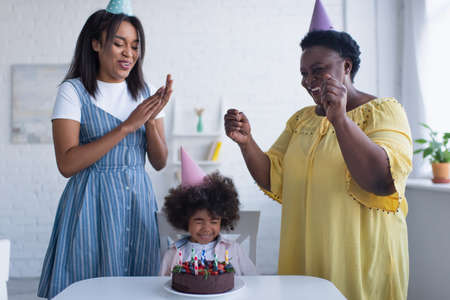 toddler african american girl with closed eyes sitting near birthday cake and mom with granny in party capsの写真素材