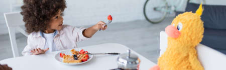 african american child proposing strawberry to toy chick during breakfast in kitchen, bannerの写真素材