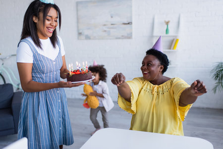 young african american woman holding birthday cake near happy mother in party capの写真素材