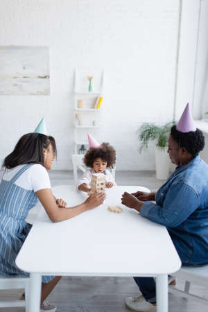 african american women and toddler girl in party caps playing wood blocks game at homeの写真素材