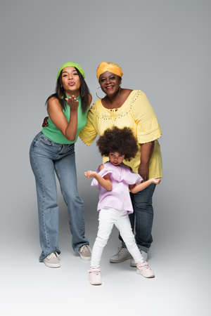stylish african american woman blowing air kiss near mother and daughter on grey