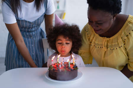 curly african american girl blowing out candles on birthday cake near granny and momの写真素材