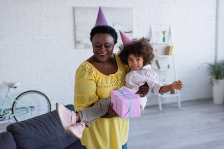 mature african american woman in party cap holding cheerful granddaughter and gift box in handsの写真素材