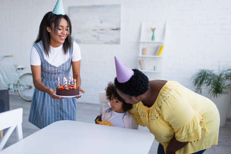 african american girl sitting at table near granny embracing her and mother holding birthday cakeの写真素材