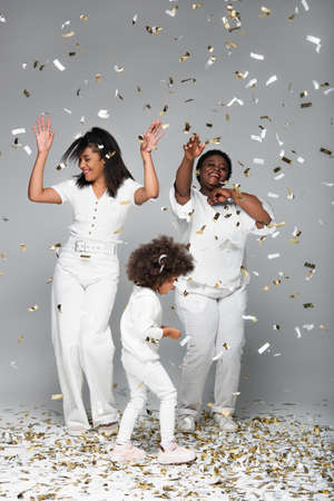happy african american women and child in white clothes under shiny confetti on greyの写真素材