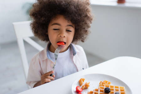 curly african american girl with closed eyes eating strawberry near tasty waffle on plateの写真素材