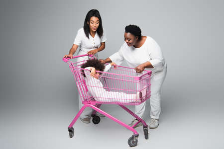 african american girl sitting in shopping trolley near happy mom and granny on greyの写真素材