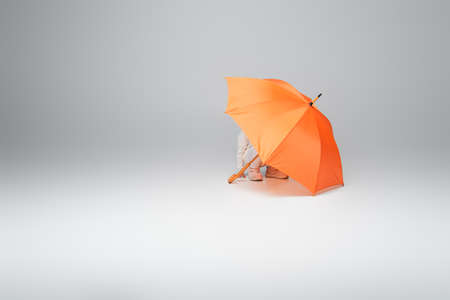 kid in white clothes hiding under orange umbrella on grey background with copy spaceの写真素材