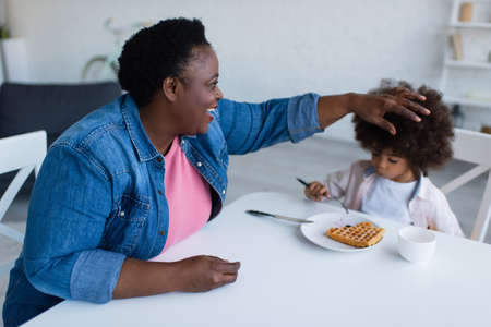 happy african american granny touching curly hair of granddaughter eating waffle for breakfastの写真素材