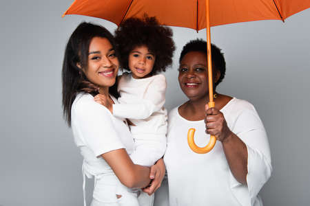 happy african american women and girl in white clothes looking at camera under orange umbrella isolated on greyの写真素材