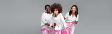 happy african american kid in shopping cart near mother and granny isolated on grey, bannerの写真素材