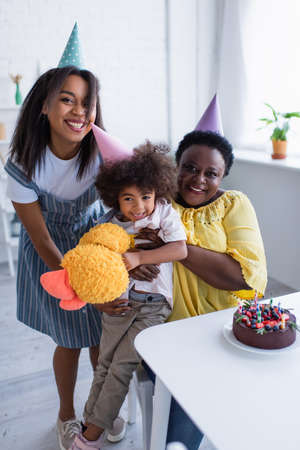 cheerful african american girl holding soft toy while mom and granny hugging her near birthday cakeの写真素材