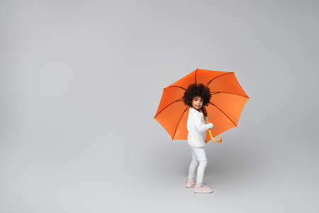 full length view of stylish african american girl under orange umbrella looking at camera on greyの写真素材