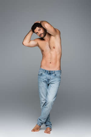 full length of good-looking and muscular man in jeans looking at camera while posing on greyの写真素材