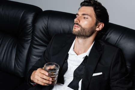 relaxed man in suit sitting on black sofa and holding glass of whiskey isolated on greyの写真素材