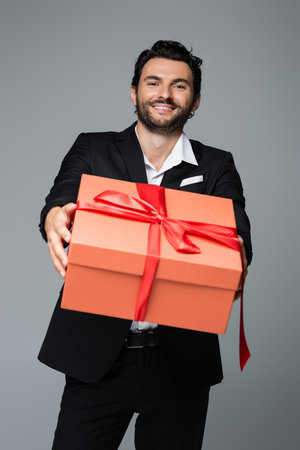 cheerful man in suit holding red gift box isolated on greyの写真素材