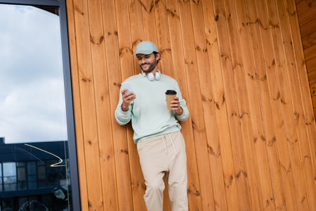 positive man in cap and wireless headphones using cellphone while holding paper cup near buildingの写真素材