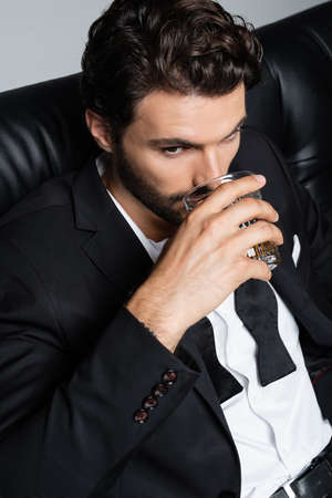 man in formal wear sitting on black sofa and drinking whiskey isolated on greyの写真素材