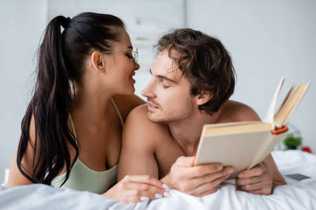 Cheerful woman in eyeglasses lying near boyfriend with book on bedの写真素材
