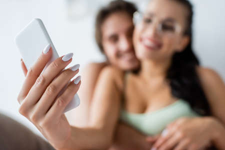 Smartphone in hand of blurred woman taking selfie near boyfriend at homeの写真素材