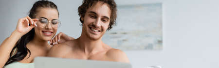 Shirtless man looking at laptop near smiling girlfriend in eyeglasses at home, bannerの写真素材