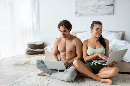 Smiling sexy couple using laptops near bed at homeの写真素材