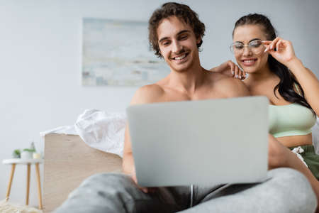 Smiling sexy woman in eyeglasses looking at blurred laptop near shirtless boyfriend in bedroomの写真素材
