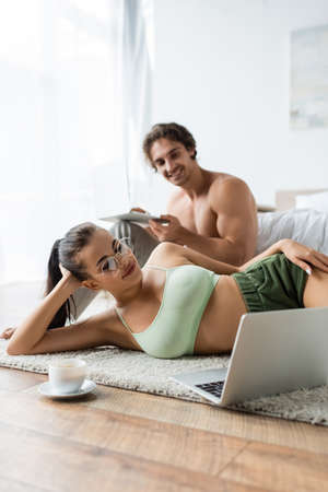 Sexy woman looking at laptop near coffee and blurred boyfriend in bedroomの写真素材