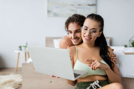 Smiling woman in top and eyeglasses pointing at laptop near shirtless boyfriend in bedroomの写真素材