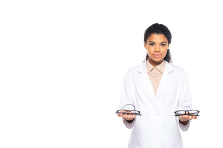Pretty african american ophthalmologist holding eyeglasses and looking at camera isolated on whiteの写真素材