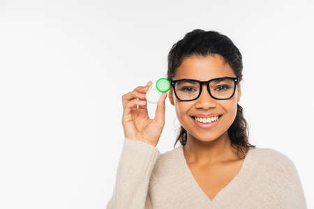 Smiling african american woman in eyeglasses holding contact lenses and looking at camera isolated...