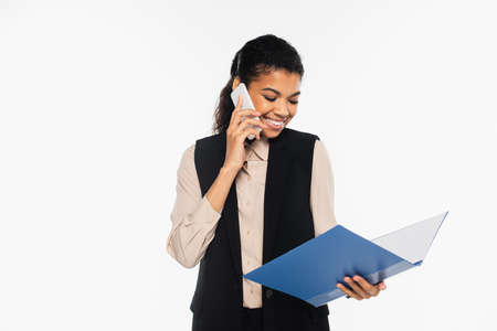 Cheerful african american businesswoman talking on mobile phone and holding paper folder isolated on whiteの写真素材