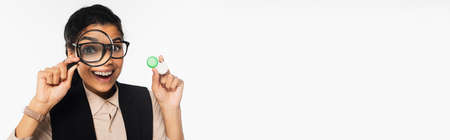 Cheerful african american businesswoman holding magnifying glass and contact lenses isolated on white, bannerの写真素材