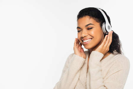 Positive african american woman in sweater listening music in headphones isolated on whiteの写真素材