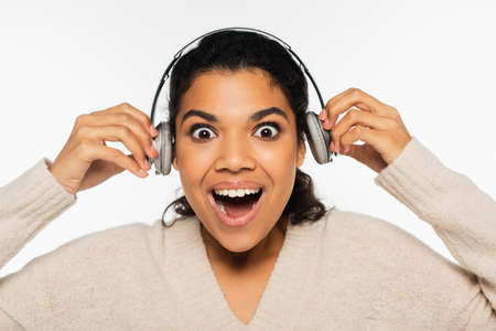 Excited african american woman in sweater holding headphones near ears isolated on whiteの写真素材