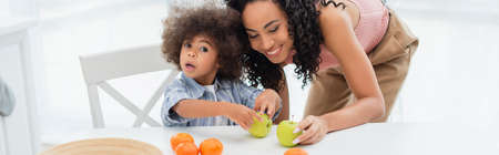 African american mom holding apple near daughter and oranges in kitchen, bannerの写真素材