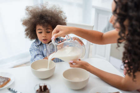 Blurred african american mother pouring milk in bowls near daughter in kitchenの写真素材
