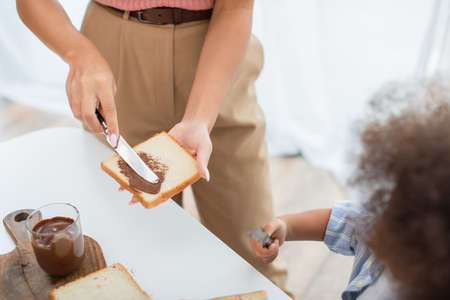 Cropped view of african american woman spreading chocolate paste on bread near blurred kidの写真素材