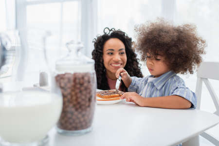 Toddler african american kid spreading chocolate paste on bread near blurred mom in kitchenの写真素材