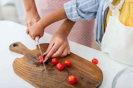 Cropped view of african american kid holding hand of mother with knife cutting cherry tomatoes in kitchenの写真素材