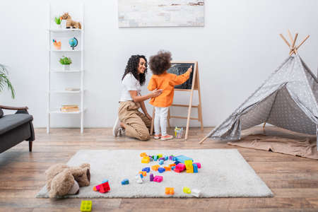 Smiling african american mom looking at kid drawing on chalkboard near building blocks and teepee at homeの写真素材