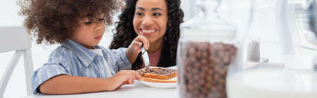 Positive african american mom looking at toddler daughter spreading chocolate paste on bread, bannerの写真素材