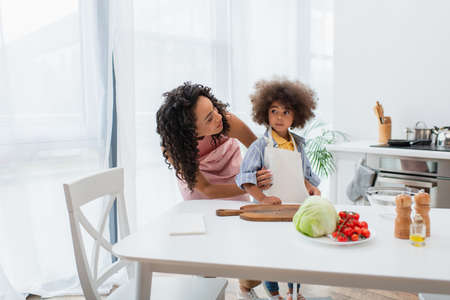 African american parent hugging daughter in apron near vegetables in kitchenの写真素材