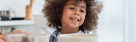 Smiling african american kid sitting near bowls and mother in kitchen, banner